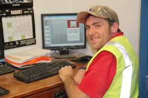 Worker at desk with computer, wearing safety vest and cap, smiling at camera.