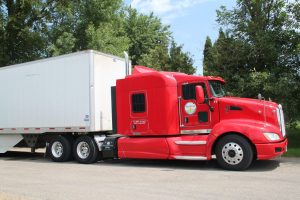 Red semi-truck with white trailer parked outdoors with trees in background.