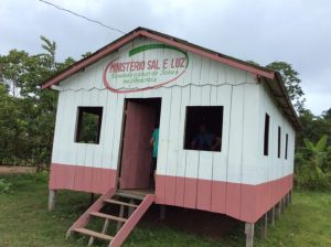 Small church building with white and pink exterior, raised on stilts, in a rural area.