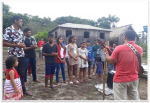 Group of people listening to a speaker outdoors in a rural setting with wooden buildings.