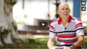 Man sitting outdoors near a tree, wearing a striped polo shirt, looking thoughtful.