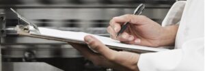 Person writing on a clipboard with a pen in a professional kitchen setting.