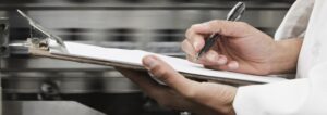 Person writing on a clipboard in a professional kitchen setting.