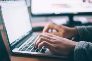 Close-up of hands typing on a laptop keyboard with a monitor in the background.